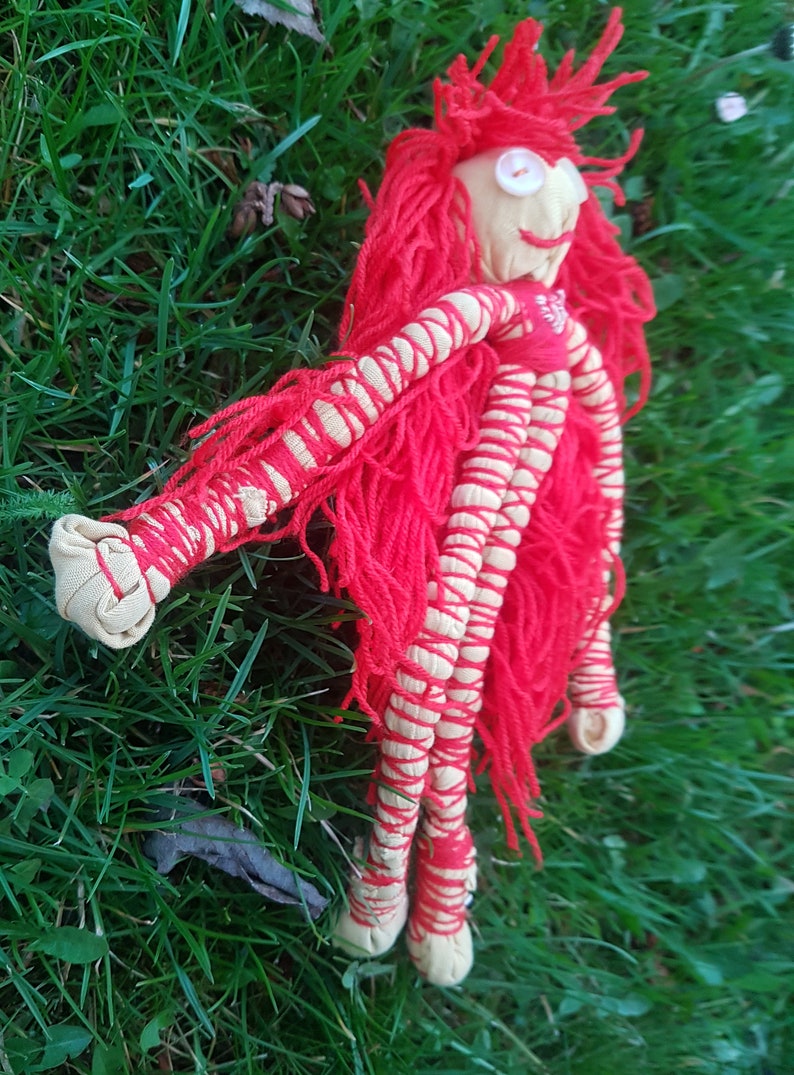May include: Handmade doll with a fabric face, button eyes, and red yarn hair. The doll's arms and legs are wrapped in cream-colored fabric and red yarn. The doll is lying on green grass.