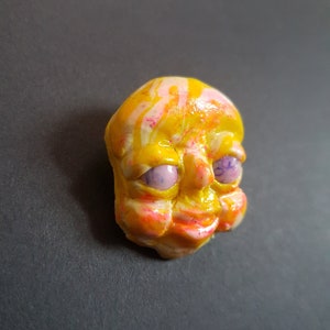 May include: A ceramic sculpture of a face, painted in yellow, pink and white with purple eyes. The sculpture is hand-painted and has a whimsical expression.