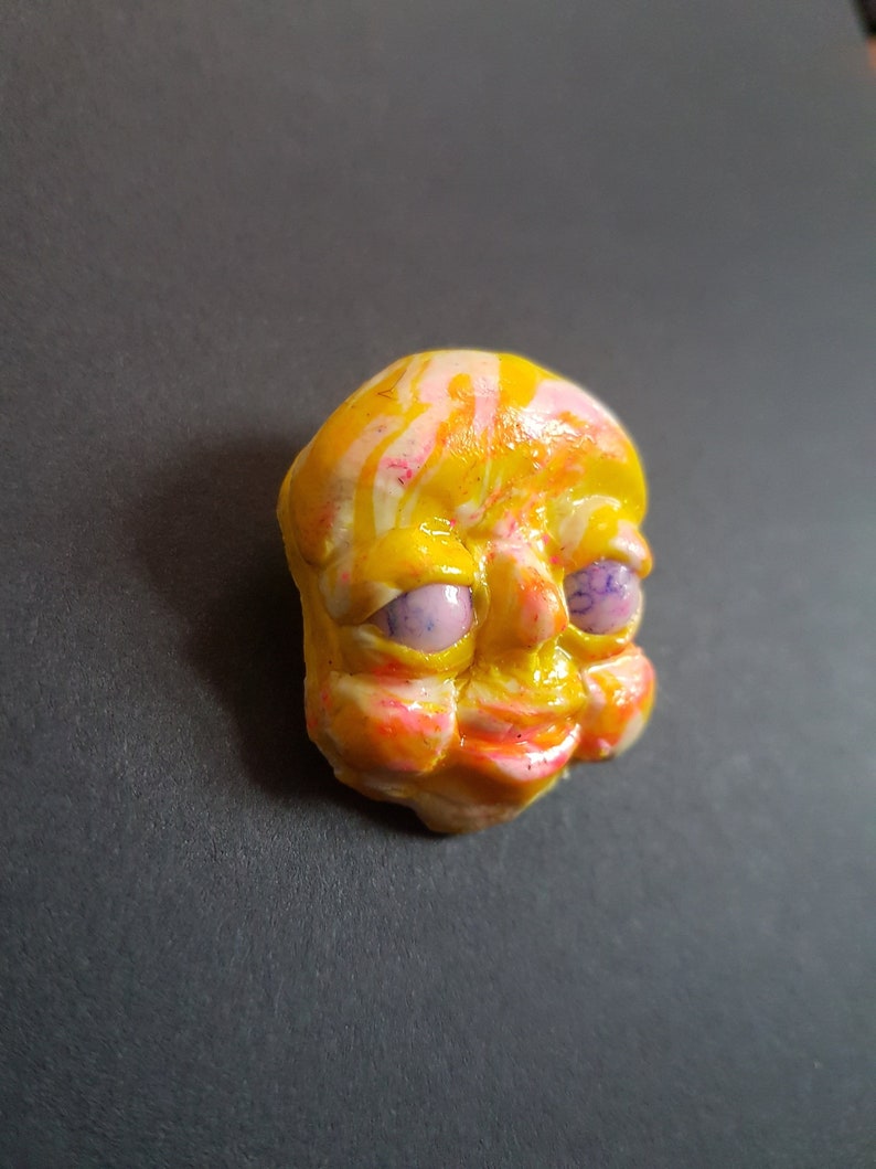 May include: A ceramic sculpture of a face, painted in yellow, pink and white with purple eyes. The sculpture is hand-painted and has a whimsical expression.