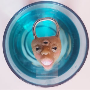 May include: A three-eyed face sculpture with a silver ring around the top of the head. The face is light brown with a pink blush and a pink mouth. The sculpture is floating in a blue liquid.