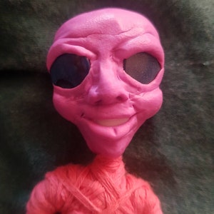 May include: A pink alien doll with black eyes and a red yarn body. The doll has a friendly expression and is smiling.