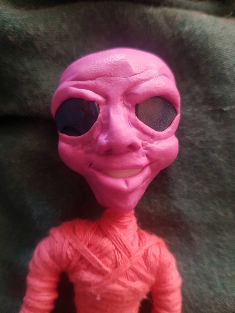 May include: A pink alien doll with black eyes and a red yarn body. The doll has a friendly expression and is smiling.