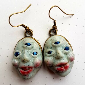 May include: A pair of earrings featuring a white ceramic face with three blue eyes and a red mouth. The earrings have a gold-toned hook.