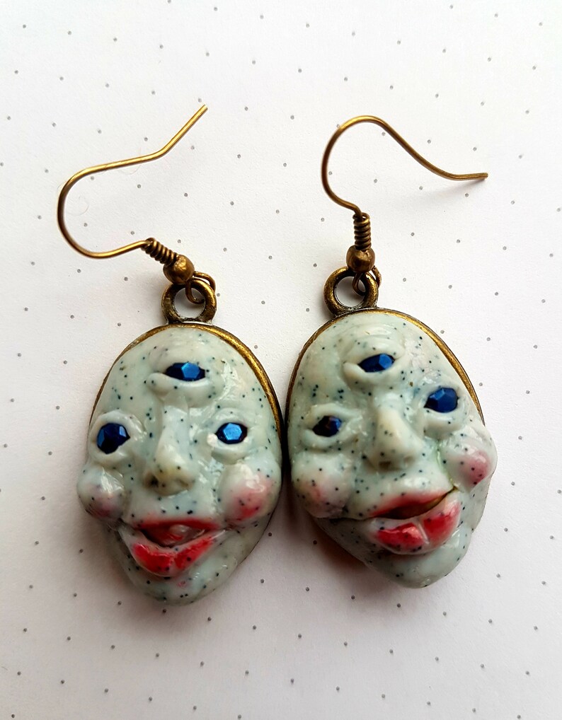 May include: A pair of earrings featuring a white ceramic face with three blue eyes and a red mouth. The earrings have a gold-toned hook.