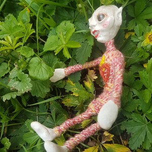 May include: A handmade doll with a white face, red and yellow yarn clothing, and a pointed ear. The doll is lying on a bed of green leaves.