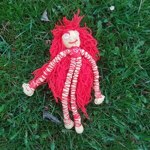 May include: A handmade doll with a tan face, red yarn hair, and red yarn limbs wrapped in tan thread. The doll has button eyes and a small red heart on its chest. The doll is lying on a bed of green grass.