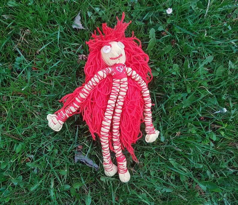 May include: A handmade doll with a tan face, red yarn hair, and red yarn limbs wrapped in tan thread. The doll has button eyes and a small red heart on its chest. The doll is lying on a bed of green grass.
