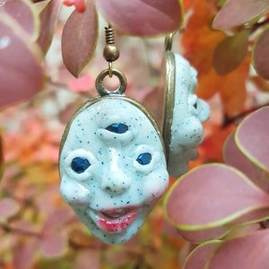 May include: A pair of earrings featuring a white, three-eyed face with blue eyes and a smiling mouth. The earrings are made of polymer clay and have a bronze finish.