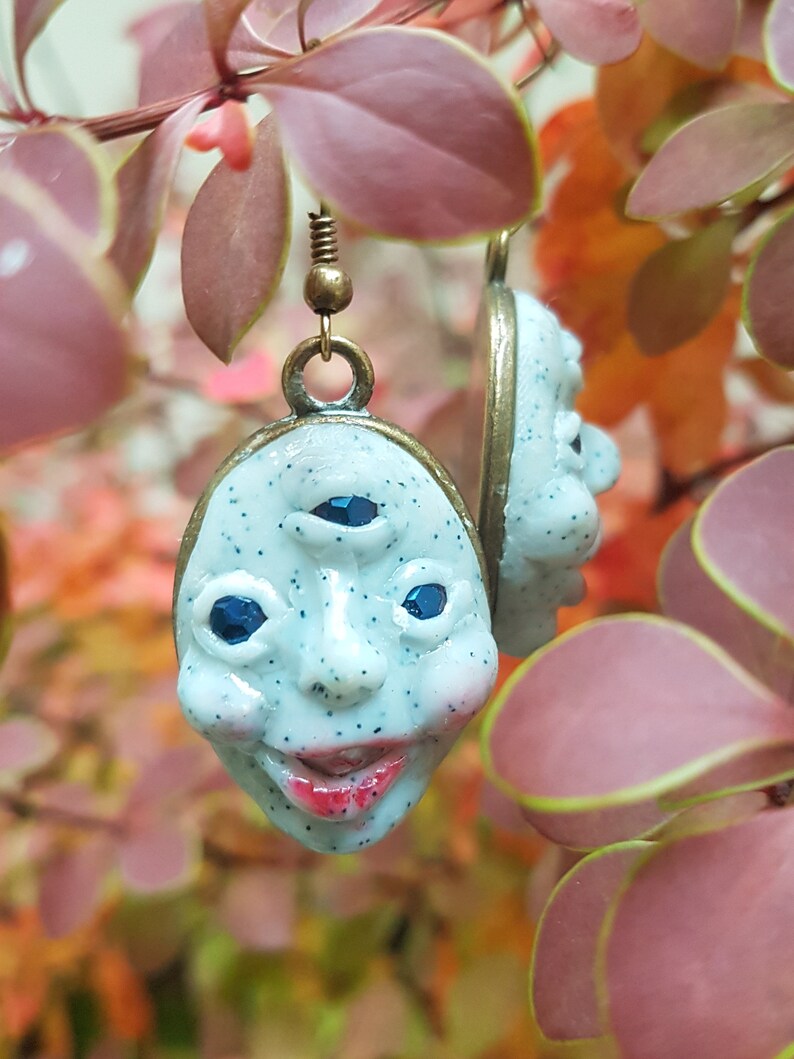 May include: A pair of earrings featuring a white, three-eyed face with blue eyes and a smiling mouth. The earrings are made of polymer clay and have a bronze finish.