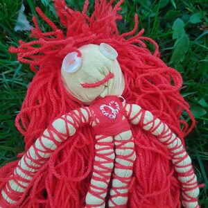 May include: Handmade doll with a yarn wig and button eyes. The doll's face is made of beige fabric with a stitched smile. The red yarn hair is long and flowing. The doll's body is red and beige with a heart design.