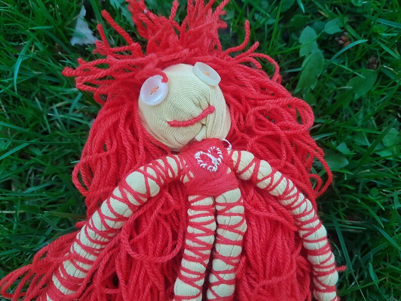 May include: Handmade doll with a yarn wig and button eyes. The doll's face is made of beige fabric with a stitched smile. The red yarn hair is long and flowing. The doll's body is red and beige with a heart design.