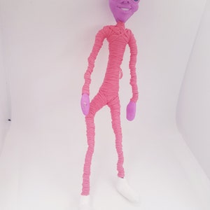 May include: A pink alien doll with a purple head and large eyes. The doll is wrapped in pink yarn and has white shoes.