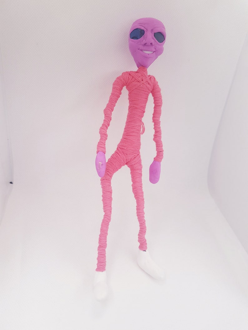 May include: A pink alien doll with a purple head and large eyes. The doll is wrapped in pink yarn and has white shoes.