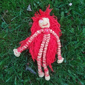 May include: A handmade doll with a cream-colored face, button eyes, and a stitched smile. The doll has bright red yarn hair and a red top with a heart design. Its arms and legs are cream-colored with red yarn detailing.