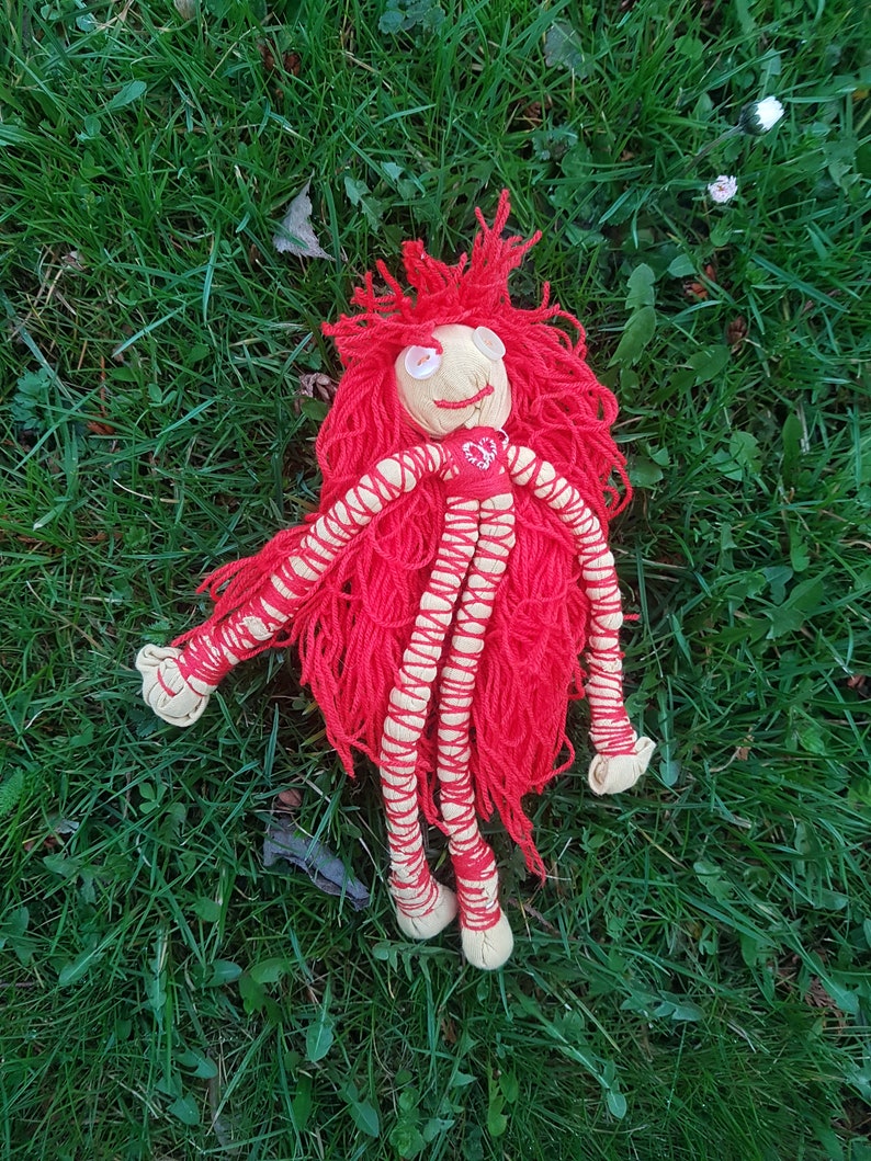 May include: A handmade doll with a cream-colored face, button eyes, and a stitched smile. The doll has bright red yarn hair and a red top with a heart design. Its arms and legs are cream-colored with red yarn detailing.