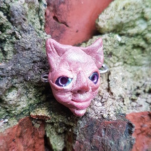 Puede incluir: Una escultura de arcilla rosa de la cara de un gato con ojos morados. La escultura est&aacute; unida a un anillo de plata.