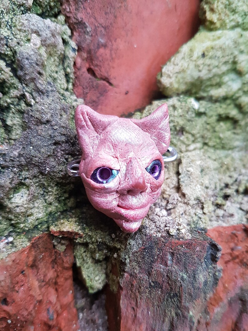 Puede incluir: Una escultura de arcilla rosa de la cara de un gato con ojos morados. La escultura est&aacute; unida a un anillo de plata.