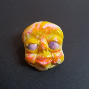 May include: A yellow, pink and white face with purple eyes. The face is made of clay and has a whimsical expression.