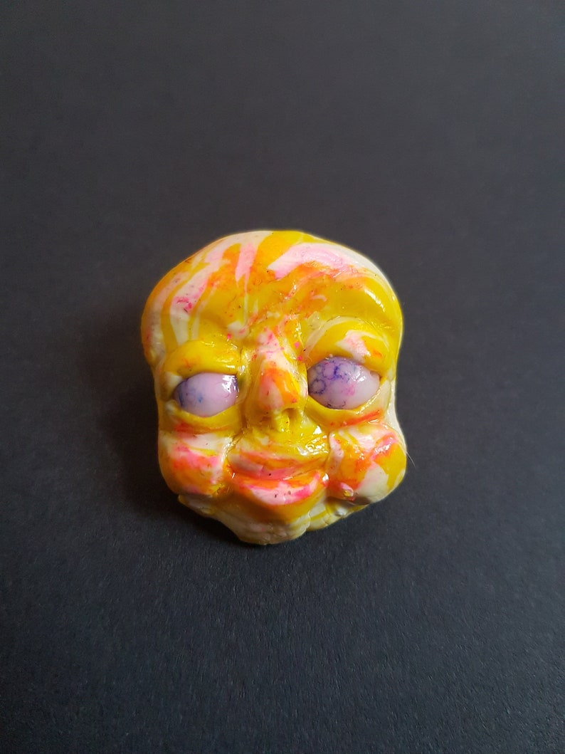 May include: A yellow, pink and white face with purple eyes. The face is made of clay and has a whimsical expression.