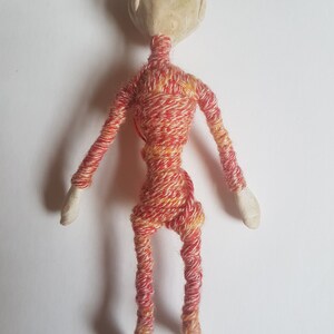 May include: A small, handmade doll with a white ceramic head and limbs wrapped in red and yellow yarn.
