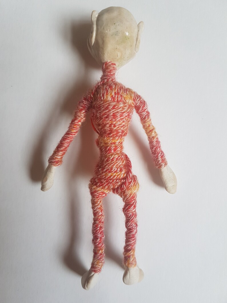 May include: A small, handmade doll with a white ceramic head and limbs wrapped in red and yellow yarn.