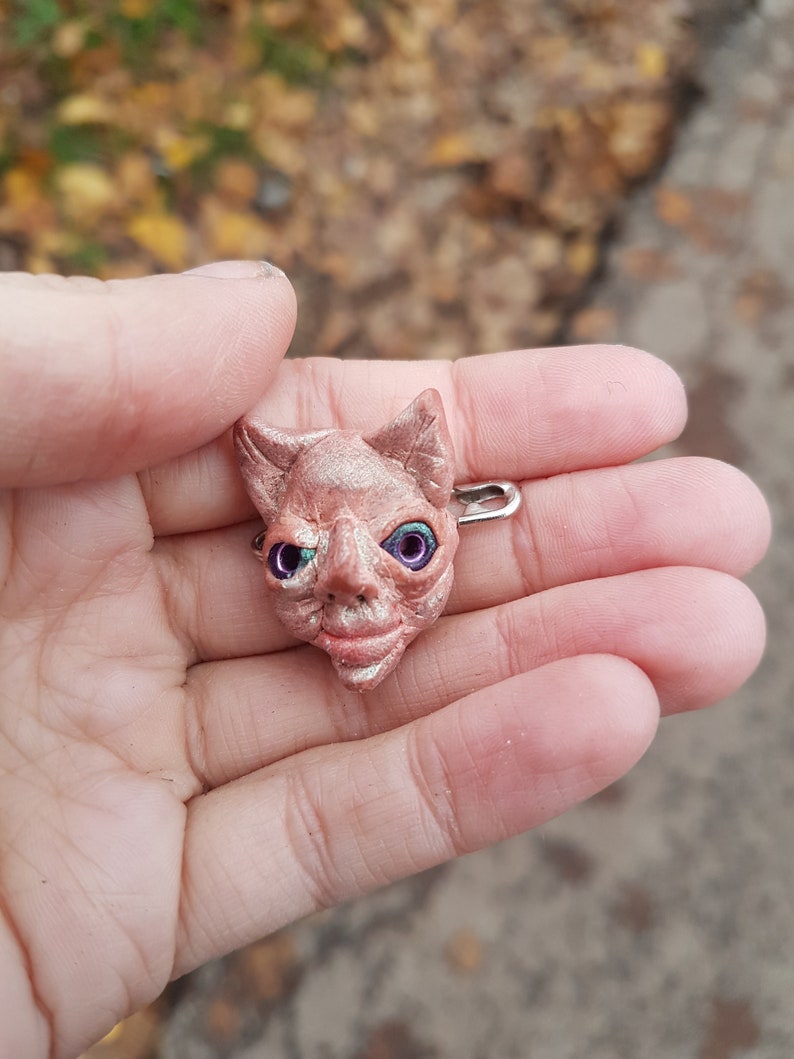 Puede incluir: Una peque&ntilde;a escultura de arcilla rosa de una cara con ojos verdes y morados. La escultura est&aacute; unida a un pasador de seguridad plateado.