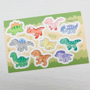 May include: A sheet of colourful dinosaur stickers with a green background. The stickers feature a variety of dinosaurs in different colours, including green, blue, red, yellow, and purple. The stickers are perfect for adding a fun and whimsical touch to any project.