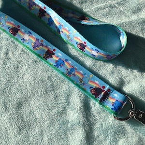 May include: A light blue lanyard with a repeating pattern of cartoon characters, rainbows, and musical notes. The lanyard has a green border and a silver-colored metal clasp.