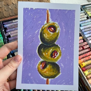 May include: An oil pastel painting of three green olives on a wooden skewer against a lilac background. The olives have red centres and are painted with thick brushstrokes. The artwork is on white paper.