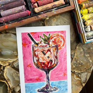 May include: An oil pastel painting of a cocktail in a stemmed glass, with a pink background and blue base. The drink is garnished with a citrus slice and mint. Oil pastels in a wooden box are visible in the background.
