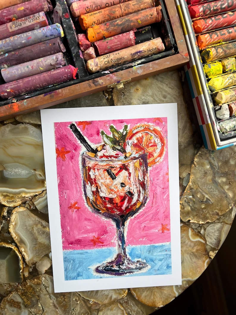 May include: An oil pastel painting of a cocktail in a stemmed glass, with a pink background and blue base. The drink is garnished with a citrus slice and mint. Oil pastels in a wooden box are visible in the background.