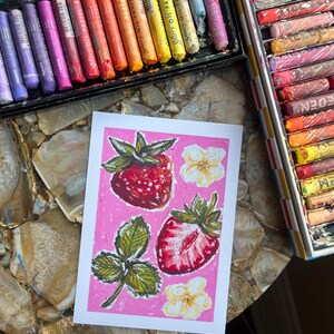 May include: A strawberry-themed artwork on pink background, featuring two whole strawberries, a sliced one, leaves, and flowers. Oil pastels in various colors are visible in open boxes, suggesting an art studio setting.