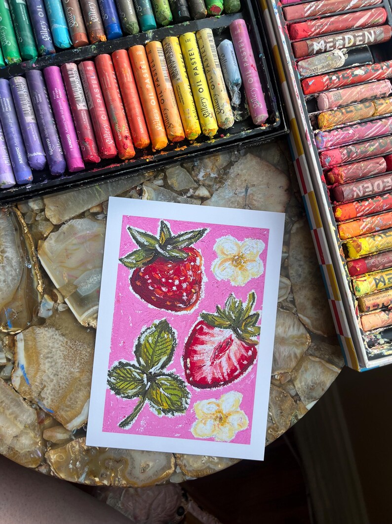 May include: A strawberry-themed artwork on pink background, featuring two whole strawberries, a sliced one, leaves, and flowers. Oil pastels in various colors are visible in open boxes, suggesting an art studio setting.