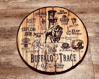 May include: A wooden barrel head with a brown stain and a black design. The design features the names of various bourbon brands, including Buffalo Trace, Old Charter, and Old Rip Van Winkle. The center of the design features a buffalo silhouette.