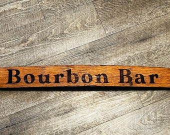 May include: A wooden sign with the words "Bourbon Bar" burnt into the surface. The sign is curved and has a dark brown stain.