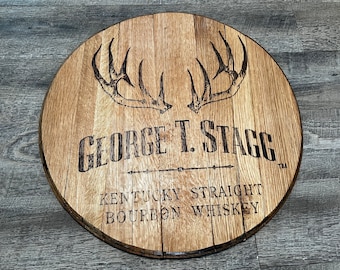 May include: A round wooden sign with a dark brown stain. The sign features a pair of stylized antlers, the text "GEORGE T. STAGG", a horizontal line with two arrowheads, and the text "KENTUCKY STRAIGHT BOURBON WHISKEY".