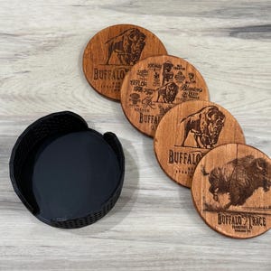 May include: A set of four round wooden coasters featuring laser-engraved buffalo imagery and Buffalo Trace distillery branding, accompanied by a black circular holder with a textured exterior finish for organized storage of the drink coasters.