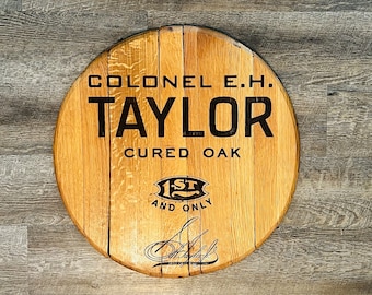 May include: A round wooden sign featuring the text "Colonel E.H. Taylor Cured Oak 1st and Only" in black lettering. The sign is crafted from reclaimed oak barrel staves, giving it a rustic look.