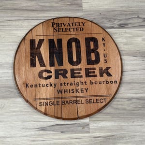 May include: Round wooden sign with the text "KNOB CREEK Kentucky straight bourbon whiskey" in black lettering. The sign also includes the text "Privately Selected" and "Single Barrel Select". The sign is made of wood and has a natural wood grain.