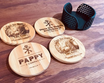 Puede incluir: Cuatro posavasos de madera con grabados, incluyendo "Pappy Van Winkle Family Reserve" y "Pappy & Company Louisville, KY." También hay un portavasos verde oscuro. Los posavasos son redondos y de madera clara.
