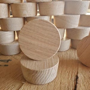 May include: A selection of unfinished wooden plugs, with two circular plugs in focus. The plugs are made of light-coloured wood, with visible grain patterns. The plugs are stacked and arranged on a wooden surface.
