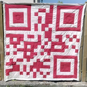 May include: A white and red quilt with a large QR code design. The quilt is made of squares of fabric sewn together.
