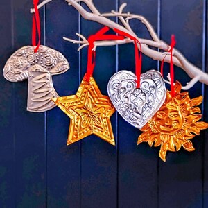 May include: Four embossed metal ornaments hanging from a white branch by red ribbons. The set includes a silver mushroom, a gold star, a silver heart with intricate scrollwork, and a gold sun face. Each piece features a textured, metallic finish.