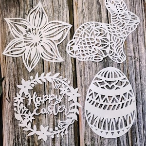 May include: White laser-cut Easter decorations on a wooden surface. The set includes a flower, a bunny, an egg, and a wreath with the text "Happy Easter". The designs have intricate cut-out patterns.