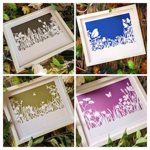 Puede incluir: Cuatro obras de arte en papel recortado enmarcadas. Cada una presenta una silueta blanca de plantas y flores sobre un fondo de color: oliva oscuro, azul, oliva claro y morado. Los marcos son blancos, sobre una superficie de madera.