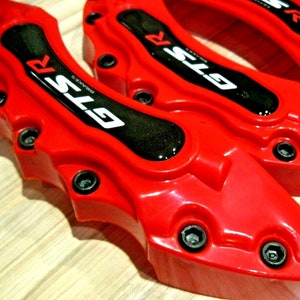 Puede incluir: Primer plano de pinzas de freno GTS rojas brillantes con detalles negros y pernos plateados. Las pinzas tienen el texto "GTS BRAKES" en blanco. La imagen muestra el diseño y la construcción de las pinzas.