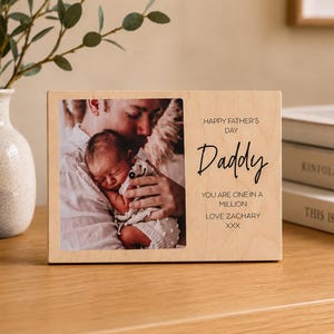 May include: A rectangular light-coloured wooden photo frame featuring a printed photograph of a person holding a newborn baby. The right side displays black text reading: HAPPY FATHER'S DAY, Daddy, YOU ARE ONE IN A MILLION, LOVE ZACHARY, XXX.