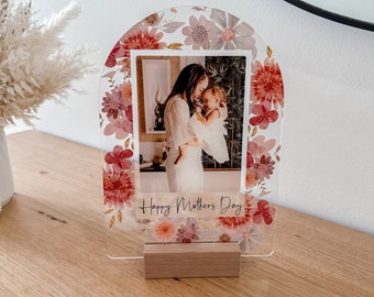 May include: A clear acrylic photo frame with a floral design and the text "Happy Mother's Day". The frame features a photo of a woman holding a child. The frame has a wooden stand.