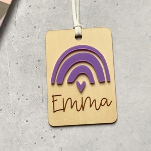 Puede incluir: Una etiqueta de madera con un arco&iacute;ris morado y el nombre "Emma" grabado en ella. La etiqueta tiene una cuerda blanca atada.