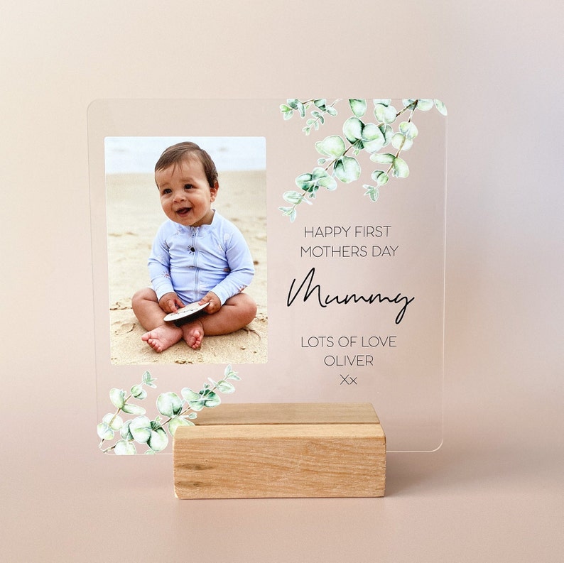 Puede incluir: Un marco de fotos de acr&iacute;lico transparente con un soporte de madera. El marco presenta una foto de un beb&eacute; sentado en una playa. El texto "Happy First Mothers Day Mummy Lots of Love Oliver xx" est&aacute; impreso en el marco.